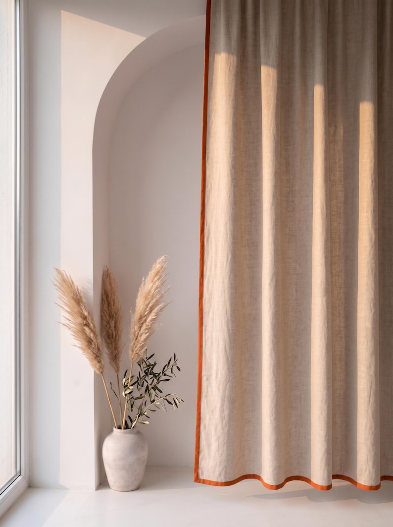 Op de afbeelding: Een kamerhoog beige gordijn met een feloranje randafwerking dat verticaal hangt. Naast het gordijn staat een lichtgekleurde keramische vaas met een verzameling hoge, vederachtige pampasgraspluimen en een takje met groene olijfbladeren.