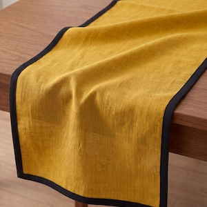 Pode incluir: Um caminho de mesa retangular com um centro de tecido amarelo-mostarda com uma textura tecida visível. O caminho de mesa é rematado com uma borda de tecido preto contrastante que emoldura os quatro lados, criando uma forma bem definida.