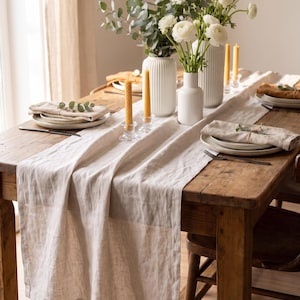 May include: A long beige linen table runner draped over a rustic wooden table, featuring white ceramic vases filled with white ranunculus flowers and eucalyptus, alongside yellow beeswax candles in glass holders and folded linen napkins.