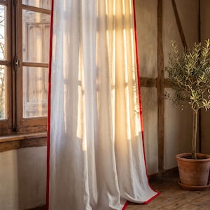 Op de afbeelding: Een kamerhoog, gebroken wit gordijn met een opvallende, effen rode rand langs de verticale zijden en de onderzoom. De stof heeft een zichtbare, licht getextureerde weving waardoor zacht, warm zonlicht door het materiaal heen kan schijnen.