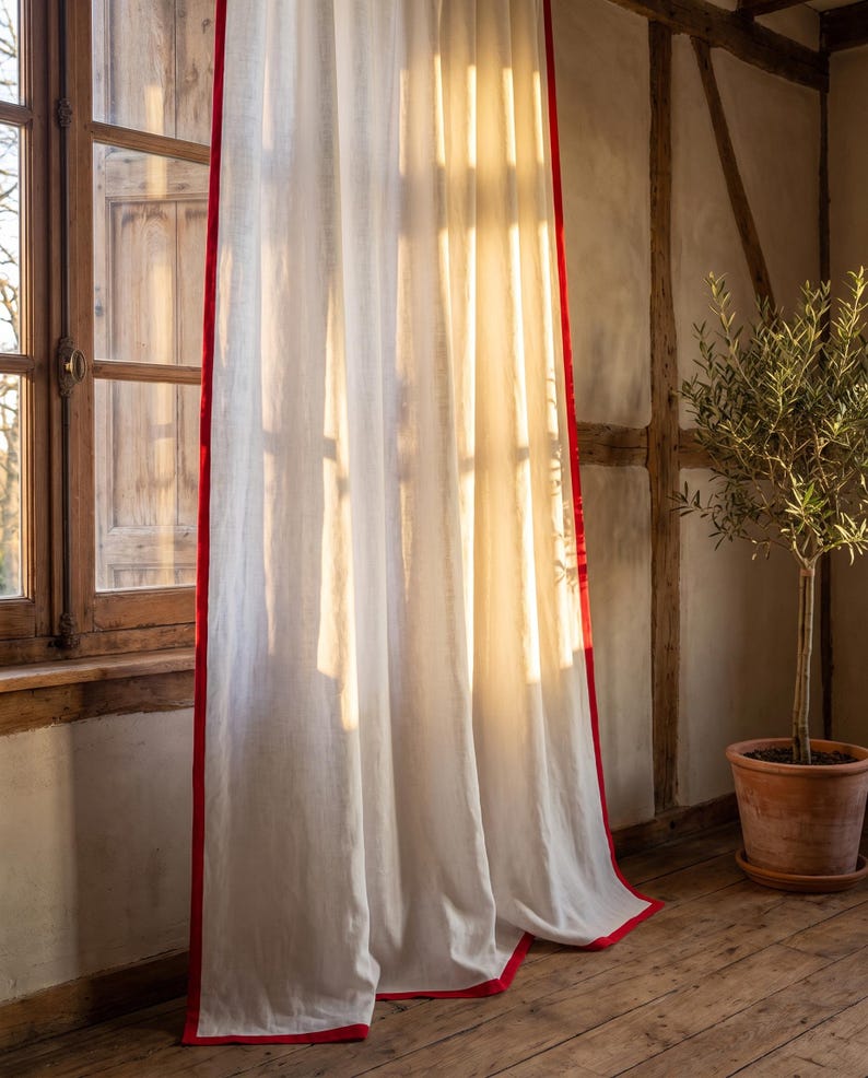 Op de afbeelding: Een kamerhoog, gebroken wit gordijn met een opvallende, effen rode rand langs de verticale zijden en de onderzoom. De stof heeft een zichtbare, licht getextureerde weving waardoor zacht, warm zonlicht door het materiaal heen kan schijnen.
