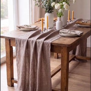 Può includere: Un tavolo da pranzo in legno rustico coperto da un lungo runner in lino color malva. Il tavolo è apparecchiato con piatti in ceramica bianca, tovaglioli di lino beige piegati, portacandele in vetro trasparente e vasi bianchi.