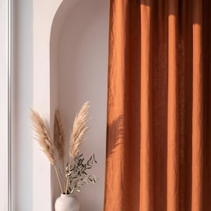 Op de afbeelding: Een kamerhoog gordijn in een effen roestoranje kleur met een zichtbare textuur. Naast het gordijn staat een lichtgekleurde, gladde, ronde vaas met daarin verschillende hoge, vederachtige pampasgraspluimen en een kleine groene olijftak.