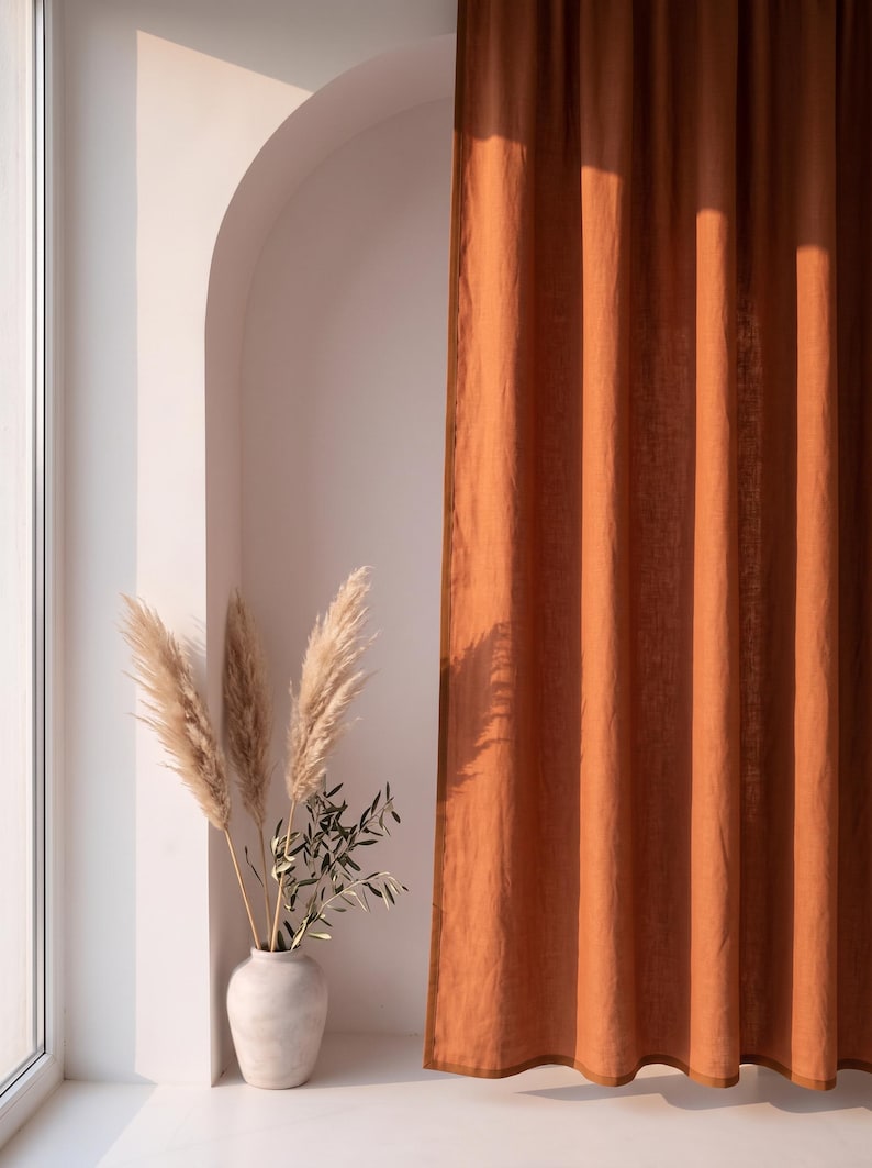 Op de afbeelding: Een kamerhoog gordijn in een effen roestoranje kleur met een zichtbare textuur. Naast het gordijn staat een lichtgekleurde, gladde, ronde vaas met daarin verschillende hoge, vederachtige pampasgraspluimen en een kleine groene olijftak.