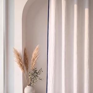 Op de afbeelding: Een kamerhoog wit gordijn met een donkerblauwe rand langs de zijkanten. De stof heeft een subtiele, textuurrijke uitstraling en hangt in verticale plooien. Naast het gordijn staat een lichtgekleurde vaas met gedroogd pampasgras en olijftakken.