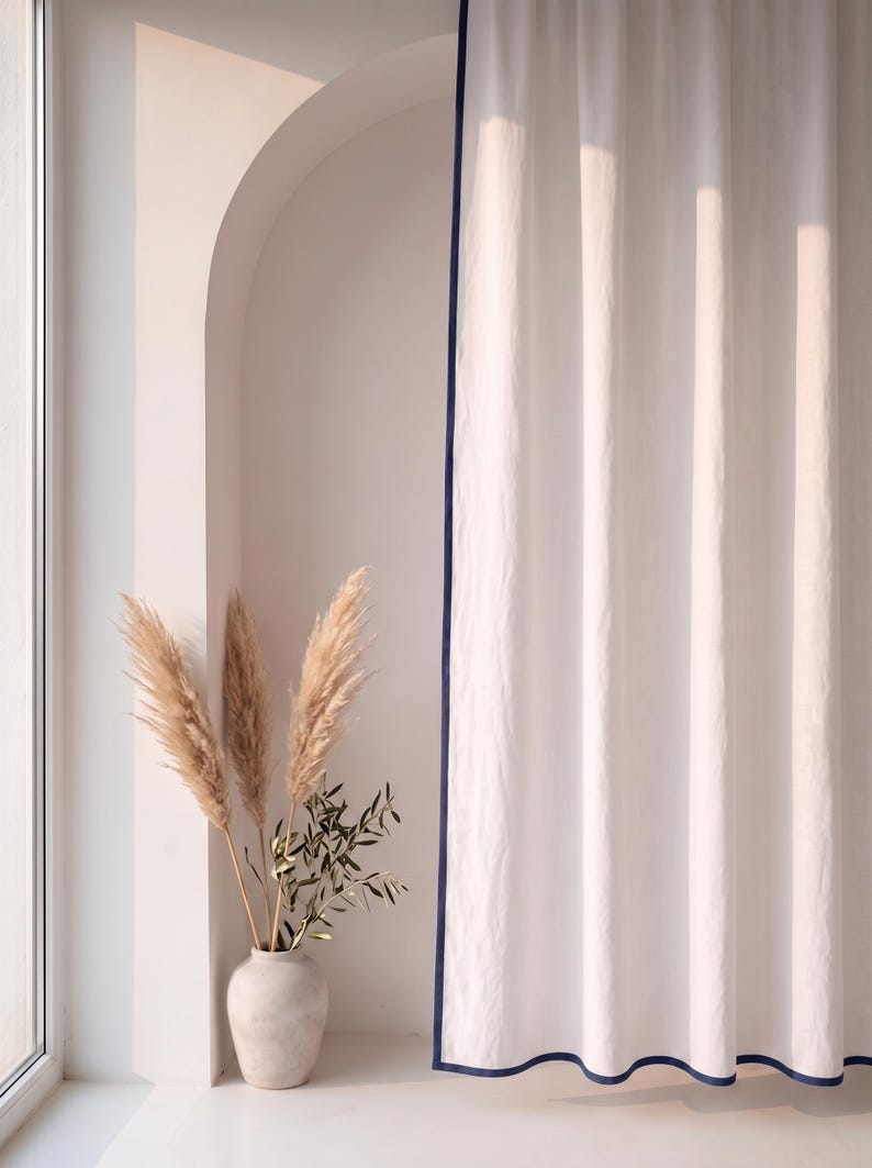 Op de afbeelding: Een kamerhoog wit gordijn met een donkerblauwe rand langs de zijkanten. De stof heeft een subtiele, textuurrijke uitstraling en hangt in verticale plooien. Naast het gordijn staat een lichtgekleurde vaas met gedroogd pampasgras en olijftakken.