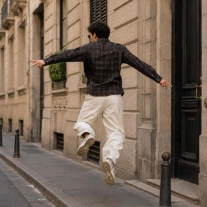 May include: A long-sleeved button-down shirt featuring a dark grey and black plaid pattern, paired with loose-fitting off-white trousers and light-colored athletic sneakers. The shirt has a classic collar and buttoned cuffs, while the pants are plain.