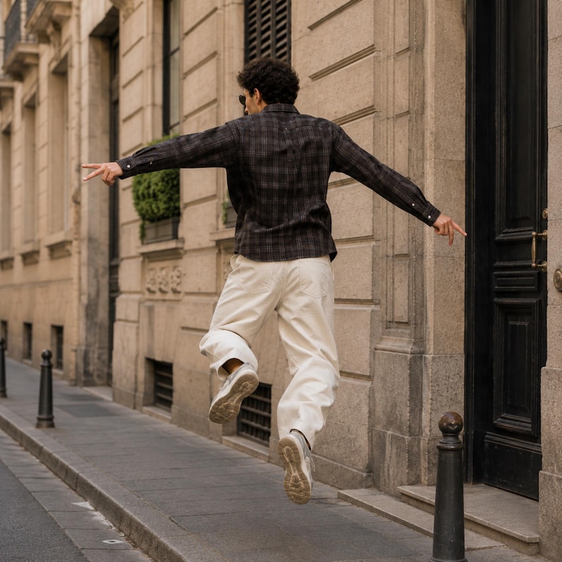 May include: A long-sleeved button-down shirt featuring a dark grey and black plaid pattern, paired with loose-fitting off-white trousers and light-colored athletic sneakers. The shirt has a classic collar and buttoned cuffs, while the pants are plain.