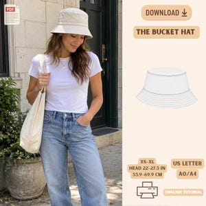 May include: A white bucket hat with visible stitching, shown as a digital pattern download. The text includes DOWNLOAD, THE BUCKET HAT, XS-XL, HEAD 22-27.5 IN, 55.9-69.9 CM, US LETTER, A0/A4, and ENGLISH TUTORIAL. The hat has a flat top and brim.