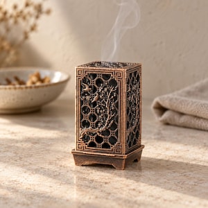 May include: A rectangular bronze-colored metal incense burner featuring an intricate lattice pattern and a relief design of birds perched on flowering branches. A thin wisp of white smoke rises from the top of the hollow, open-work structure.