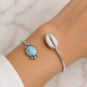 May include: A silver cuff bracelet featuring two decorative elements. One side has a light blue oval stone with silver accents, and the other side has a white seashell. The bracelet is worn on a wrist.