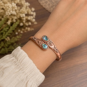 May include: A copper-toned metal cuff bracelet featuring an open-ended design with two teardrop-shaped iridescent abalone shell inlays. The metal band displays intricate beaded and twisted wire detailing along the edges of the setting.