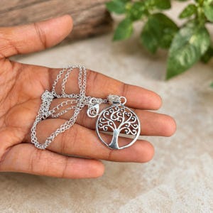 May include: A silver-toned necklace featuring a circular pendant with an intricate cutout design of a tree with spreading branches and roots. The pendant is attached to a delicate, thin silver-colored metal chain with small, uniform circular links.