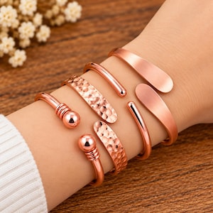 May include: A set of four rose gold-toned metal cuff bracelets. Two bracelets feature a smooth, polished finish with rounded ends, while the other two bracelets display a textured, hammered surface with spherical accents at the openings.