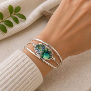 May include: A silver bracelet with an oval abalone shell centerpiece. The shell features iridescent blues, greens, and purples. The bracelet has a simple, elegant design with a polished finish. The bracelet is worn on a wrist.