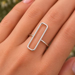 May include: A silver ring featuring a rectangular open design. The ring is a minimalist style, with a thin band and a long, slender rectangle as the focal point. The ring is shown on a hand.