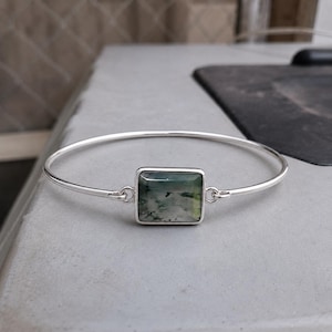 May include: A silver bangle bracelet featuring a central rectangular moss agate gemstone. The stone displays translucent white and green hues with dark, dendritic inclusions. The metal band is thin and polished, connecting to the stone setting.