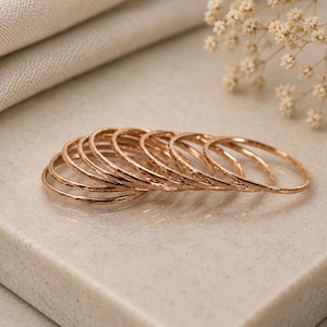 Peut inclure: Un ensemble de sept bagues fines en métal de couleur or rose avec une finition martelée et texturée. Les bagues sont empilées légèrement décalées les unes des autres, mettant en valeur leur forme circulaire et leur surface métallique réfléchissante.