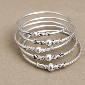 May include: Five silver bangles are stacked, each with a central silver bead and intricate wire detailing. The bangles are presented on a neutral surface, highlighting their simple and elegant design. The bangles are approximately 6 cm in diameter.
