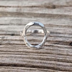 Puede incluir: Un anillo de plata con un diseño ovalado. El anillo tiene una textura martillada y está colocado sobre una superficie de madera. El centro abierto del anillo es el punto focal, destacando su forma y estilo únicos.