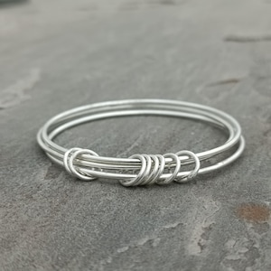 May include: A set of three silver bangles. The bangles are connected with wrapped wire detailing. The jewellery is displayed on a grey surface.