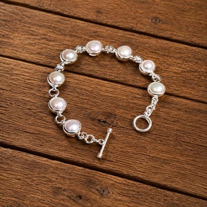 May include: A silver chain bracelet featuring eight round, white, lustrous pearls set in individual silver bezels. The bracelet is secured with a silver toggle clasp consisting of a circular ring and a T-shaped bar, creating a classic link design.