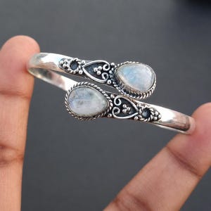 May include: A silver-toned cuff bracelet featuring two pear-shaped, iridescent moonstone cabochons. The bracelet has an open design with ornate, swirling details around the stones. The bracelet is held by a hand.