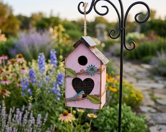 May include: A pink wooden birdhouse with a heart-shaped aperture and a small blue bird. The birdhouse is decorated with faux vines, leaves, and a blue flower. It hangs from a black metal shepherd's hook in a garden setting.