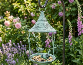 May include: A verdigris-coloured bird feeder with a conical roof and leaf detailing, suspended from a curved metal hook. The feeder has two bird figurines and a tray filled with birdseed. The background shows a garden with pink and purple flowers.