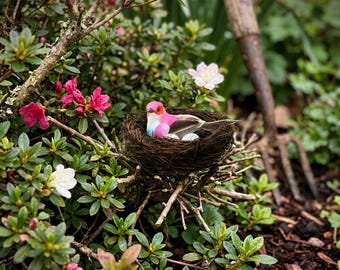 May include: A colourful bird figurine with pink, blue, and brown plumage rests in a brown nest containing two speckled eggs. The nest is surrounded by green leaves and pink and white flowers.