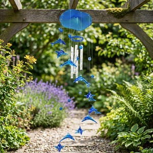 Peut inclure: Un carillon éolien bleu en forme de dauphins suspendu à l'extérieur. Il comprend un disque bleu, des dauphins et des perles de verre. Des carillons transparents sont suspendus au centre. Le fond montre un jardin verdoyant.