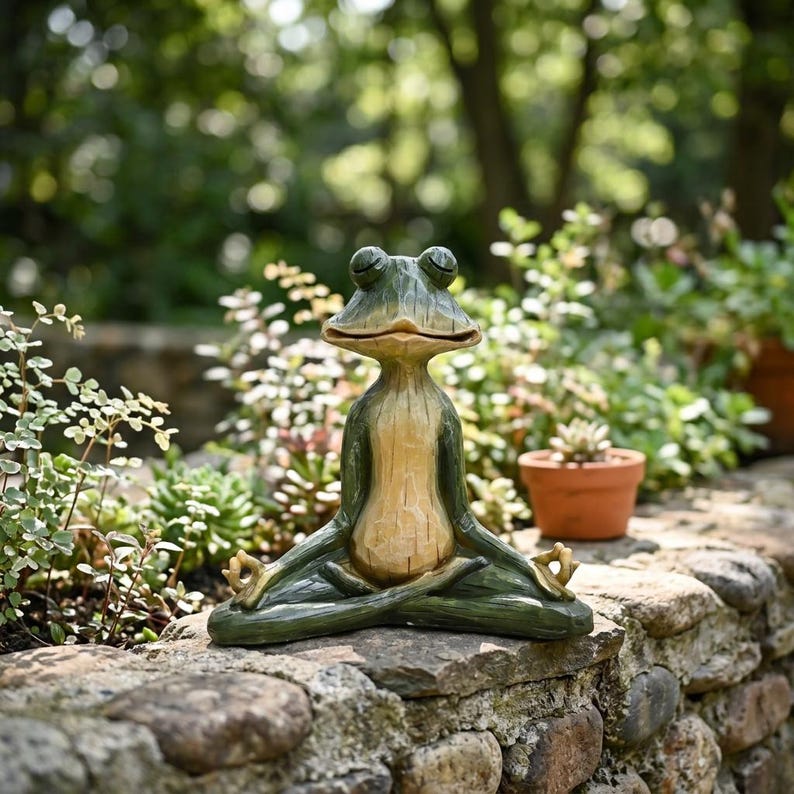 K&ouml;nnte beinhalten: Eine gr&uuml;ne und beige Froschfigur in sitzender Meditationshaltung mit geschlossenen Augen und H&auml;nden in Mudra-Position auf den Knien. Der Frosch hat ein glattes, geschnitztes Aussehen mit einem hellen Bauch sowie dunkelgr&uuml;nen Gliedma&szlig;en.