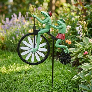 Könnte beinhalten: Ein Gartenstecker aus Metall mit zwei grünen Fröschen auf einem schwarzen Hochrad. Das große Vorderrad hat weiße, blütenblattförmige Speichen, während das kleine Hinterrad schwarz ist. Die Frösche tragen kleine rote Hüte und einer trägt eine rote Hose.