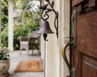 May include: A dark brown cast iron wall-mounted door bell featuring a decorative bird perched on a leafy branch above the bell. A long, thin cord with a small weight hangs from the centre of the bell, which is attached to a light-coloured door frame.