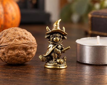 May include: A small, antique-gold coloured metal figurine of a Highland cow dressed as a witch. The cow wears a pointed hat and a cape, holds a broomstick, and stands next to a small pumpkin. The figure has a textured, detailed finish.