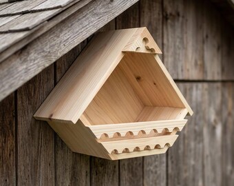 May include: A light-coloured wooden birdhouse with a triangular roof and a decorative scalloped front railing. The structure features a pointed gable top with a small cutout detail and is mounted against a weathered, vertical wood-plank wall.