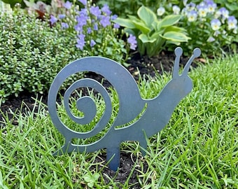 May include: A flat, grey metal garden stake shaped like a snail with a prominent spiral shell design and two antennae. The silhouette is cut from a single piece of sheet metal and features a pointed base for insertion into the ground.