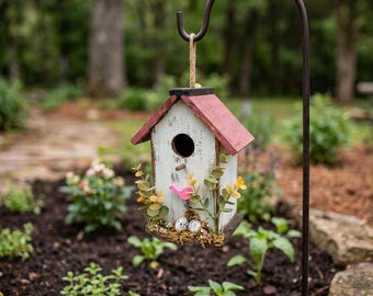 May include: A distressed white birdhouse with a red roof hangs from a black metal shepherd's hook. The birdhouse is decorated with faux greenery, a pink bird, and two speckled eggs. A charming garden ornament.