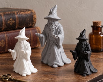 May include: Three wizard figurines in white, grey, and black, each depicted in a kneeling position with arms extended forward. The figures wear pointed hats and long robes with detailed beards. The set is arranged on a wooden surface.