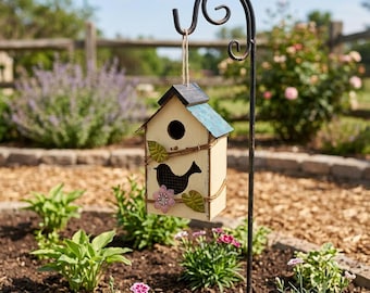 May include: A cream-coloured birdhouse with a blue-green roof and a black bird silhouette, suspended from a black metal shepherd's hook. The birdhouse is decorated with floral and leaf accents. The scene is set in a garden with plants and a wooden fence.