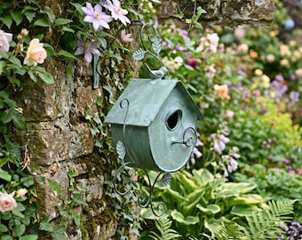May include: A light green metal birdhouse with a weathered finish, featuring a peaked roof, a circular entrance hole, and decorative scrollwork accents. A small metal bird figure sits on the roof peak, and the structure is suspended by a metal bracket.