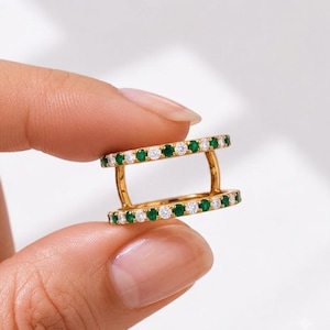 May include: A gold double-band ring featuring two parallel rows of alternating round-cut green emeralds and clear white diamonds. The two bands are connected by two vertical gold bars, creating a structured, open-frame design with a polished finish.