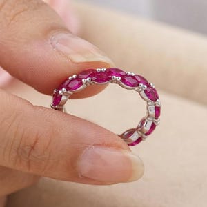 May include: A silver eternity band featuring a continuous row of marquise-cut vibrant pinkish-red gemstones set in a polished metal frame. The ring displays a repeating pattern of stones secured by small metal prongs, creating a circular design.