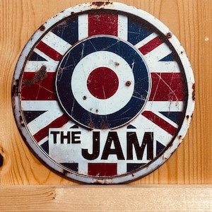 Op de afbeelding: Een rond metalen bord met een rood, wit en blauw Union Jack vlagpatroon en een centraal doelmotief. De tekst THE JAM staat in zwarte hoofdletters onderaan gedrukt. Het bord vertoont zichtbare roestplekken en krassen op het oppervlak.