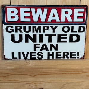 Könnte beinhalten: Ein rechteckiges Metallschild mit weißem Hintergrund und rotem Rand. Der obere Bereich zeigt das Wort BEWARE in weißer Schrift auf rotem Grund. Darunter steht in schwarzer Schrift GRUMPY OLD UNITED FAN LIVES HERE! Das Schild hat ein abgenutztes Finish.