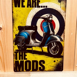 Könnte beinhalten: Ein rechteckiges Metallschild mit einem blauen Motorroller vor einem gelben Hintergrund mit einer rot-weiß-blauen Zielscheibengrafik. Der Text WE ARE... und THE MODS ist in fetten schwarzen Buchstaben oben und unten auf dem Schild aufgedruckt.