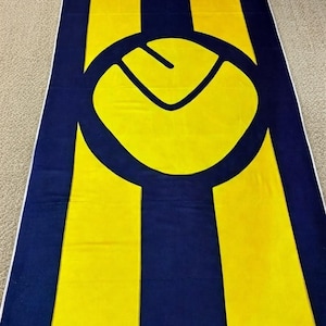 Può includere: Un telo mare rettangolare caratterizzato da un audace motivo a righe verticali blu navy e giallo brillante. Il centro del telo presenta un grande emblema circolare contenente una forma grafica gialla stilizzata su uno sfondo blu navy.