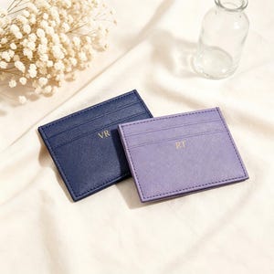 May include: Two rectangular card holders with a textured finish. One is dark navy blue with the gold embossed initials VR, and the other is light purple with the gold embossed initials RT. Both feature horizontal slots for card storage.