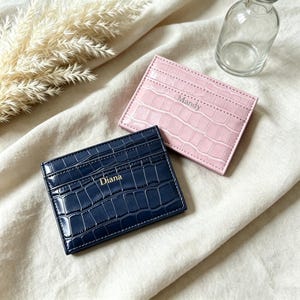 Può includere: Due portacarte rettangolari con una texture goffrata a coccodrillo. Un portacarte è rosa chiaro con il nome Mandy impresso in argento, l'altro è blu navy scuro con il nome Diana impresso in oro. Entrambi sono dotati di diverse fessure.