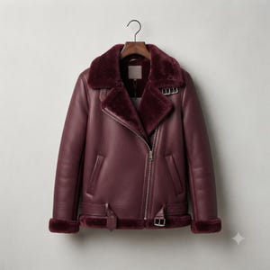 May include: A deep burgundy leather jacket featuring a thick, matching faux fur lining on the wide collar, cuffs, and hem. The jacket includes an asymmetrical silver-tone zipper closure, two side pockets, and an adjustable belt at the waist.